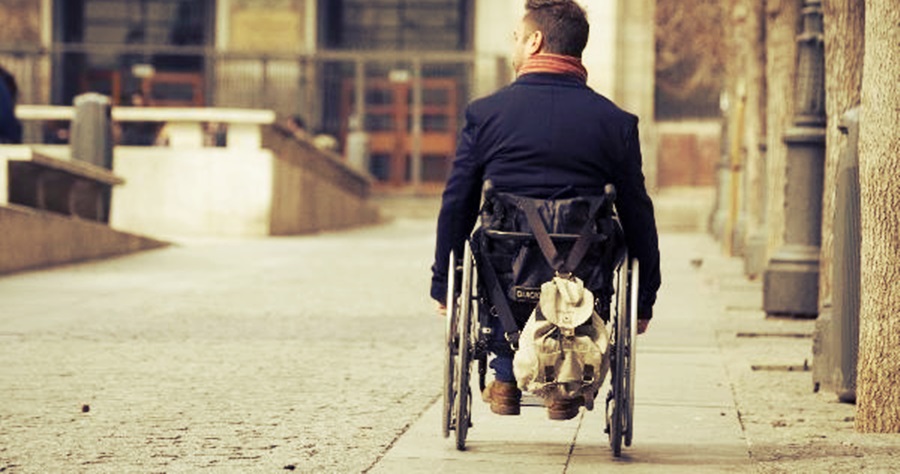 Un hombre en silla de ruedas va por la calle, se le ve de espaldas y una mochila cuelga del respaldo de su silla de ruedas