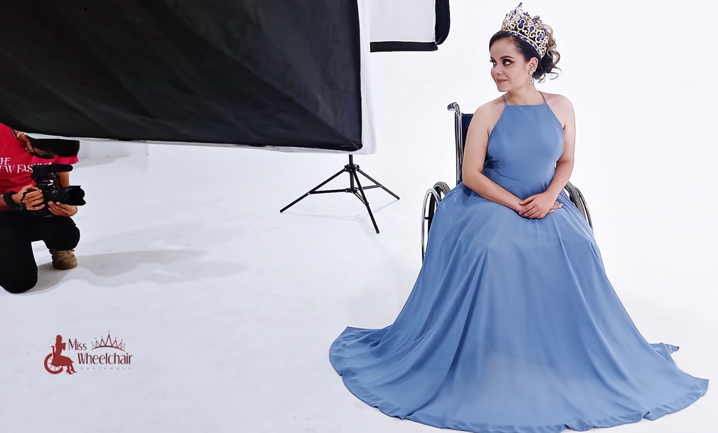 En su silla de ruedas, con su corona y vestida con un vestido de noche color azul tenue sin brazos, María posa para un fotógrafo en un estudio fotográfico color blanco. 