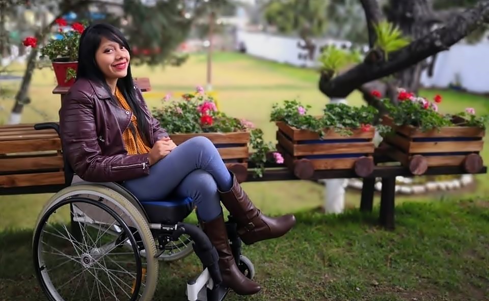 Con un parque de fondo, Pahola sonríe a cámara en su silla de ruedas y vestida de pantalón de lona con chaqueta y botas de cuero. 