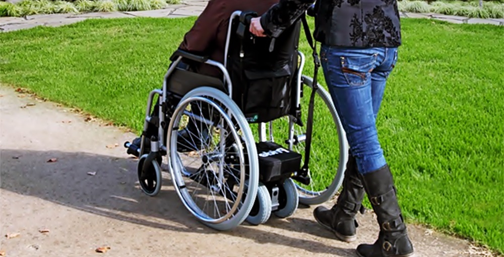 Un parsona empuja a otra en su silla de ruedas en algun parque, ambos de espaldas y medio cuerpo. 