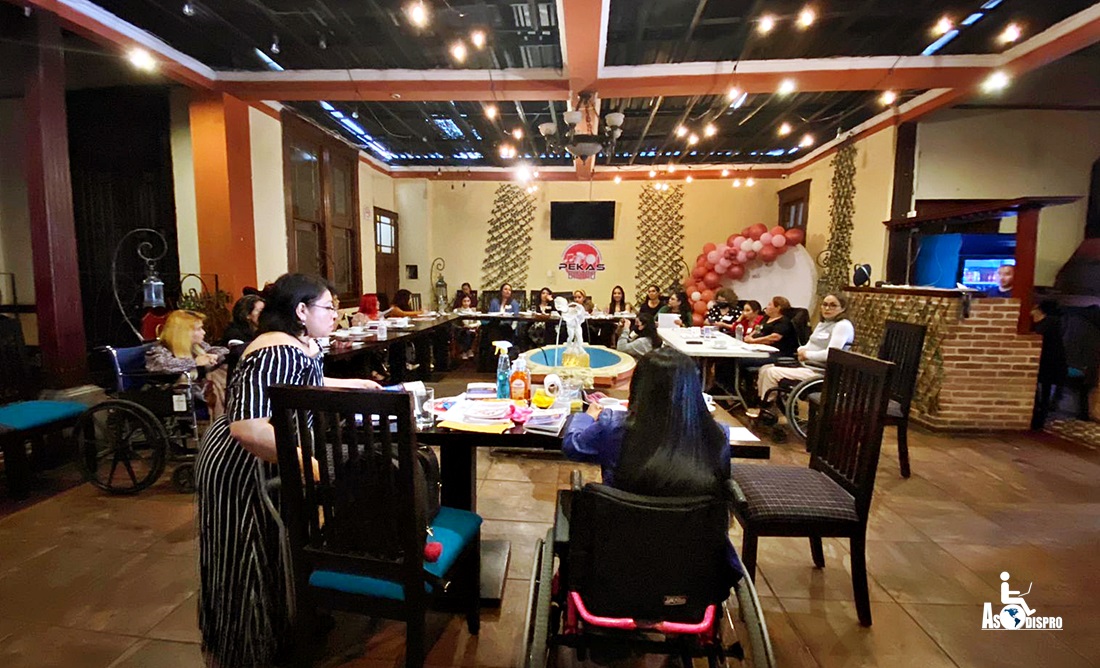 Se observa por detrás de una mesa en la que Pahola Solano se dirige a un auditorio de mujeres con discapacidad ubicadas en mesas alrededor. 