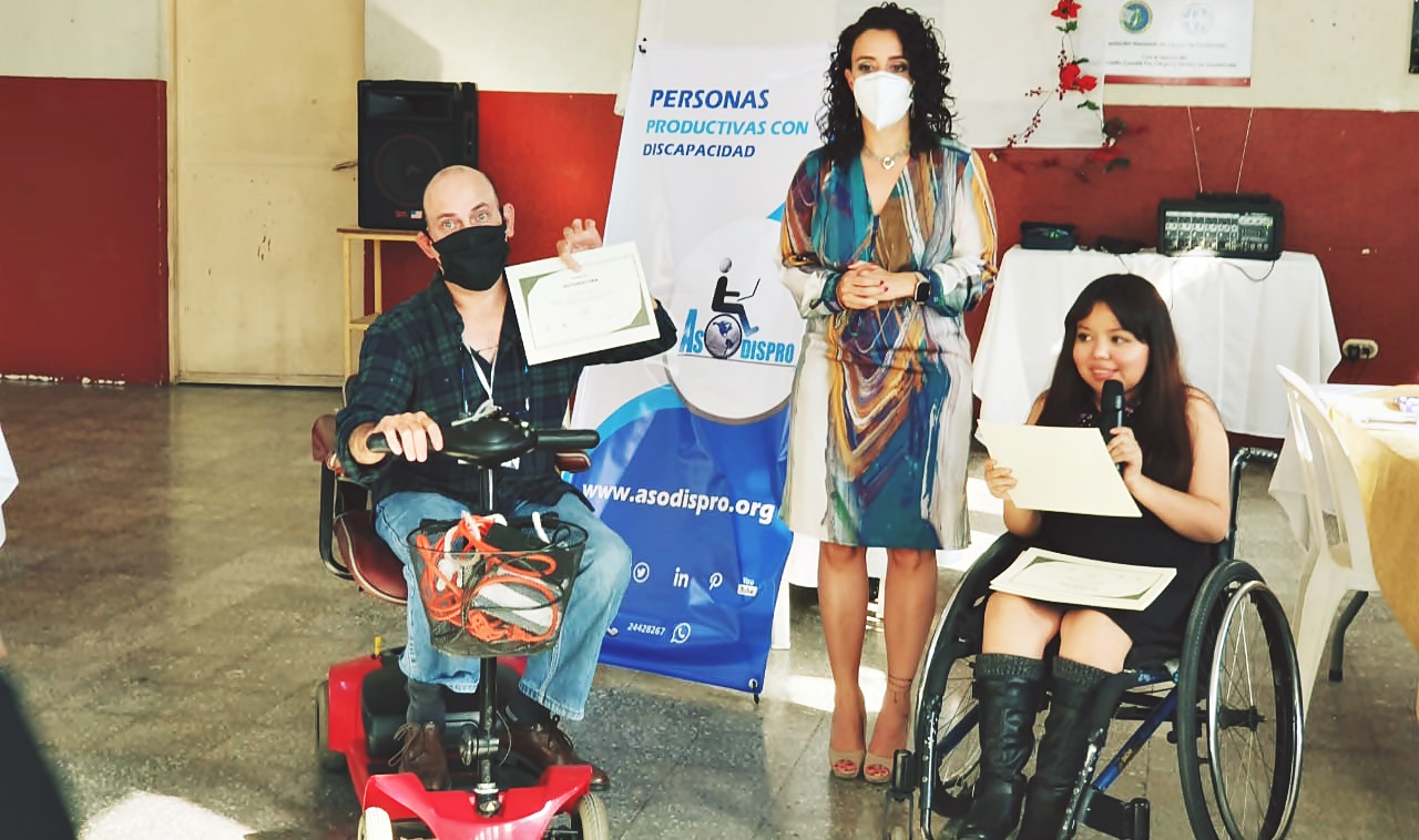 Herbert Moll, en su silla de ruedas eléctrica, sostiene su diploma de participación en el taller, la Licda. Eimy Soto está parada a su lado, mientras que Jesica Gómez, en su silla de ruedas y con el micrófono, prepara otro diploma para ser entregado. 