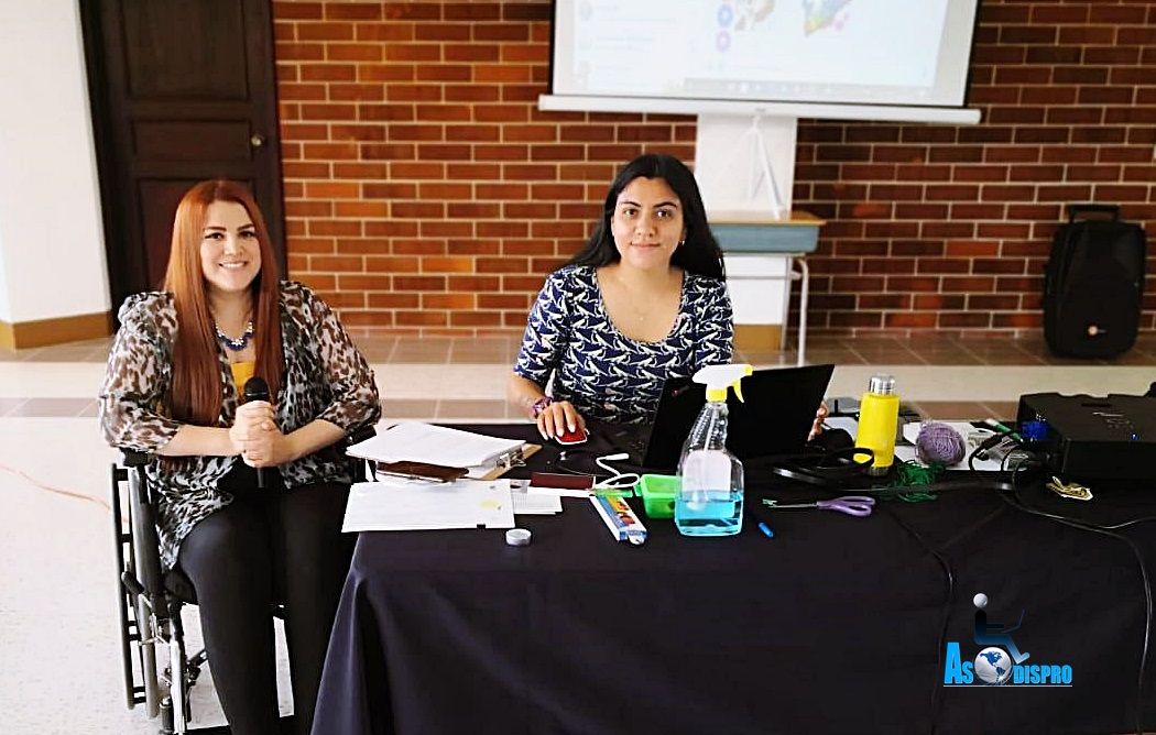 Lucrecia en su silla de ruedas y Raquel sentada a su lado ven a cámara desde la mesa donde dirigieron el evento.