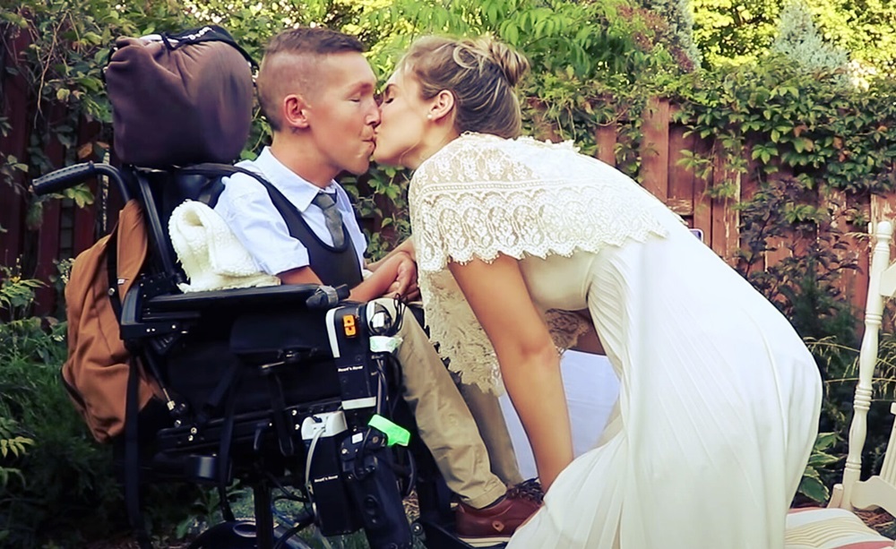 En un jardín, Hannah Aylward y Shane se besan. Ella viste un vestido blanco con encajes y él está con una camisa de cuello, un chaleco con corbata. 