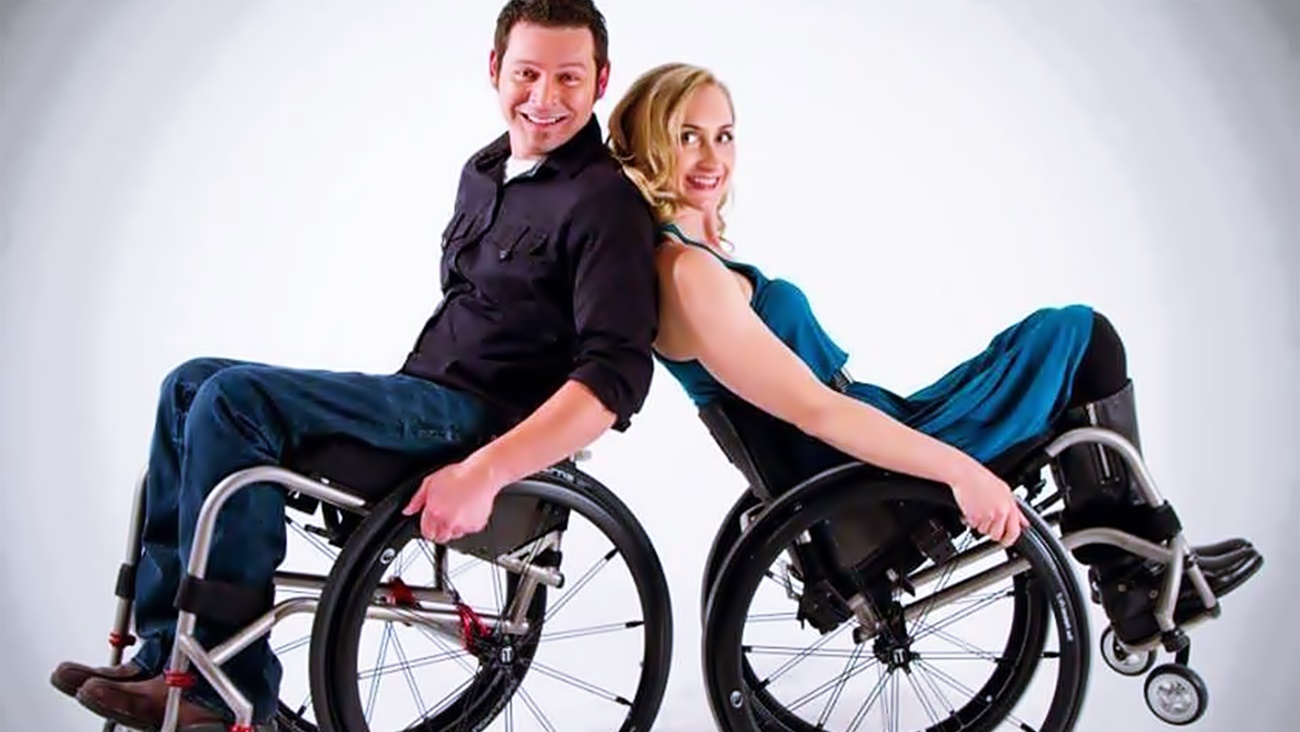 Dos personas, un hombre y una mujer, están en sus sillas de ruedas, de espaldas uno con el otro, recostados ligeramente y levantando la parte delantera de las sillas, ambos sonríen a cámara.