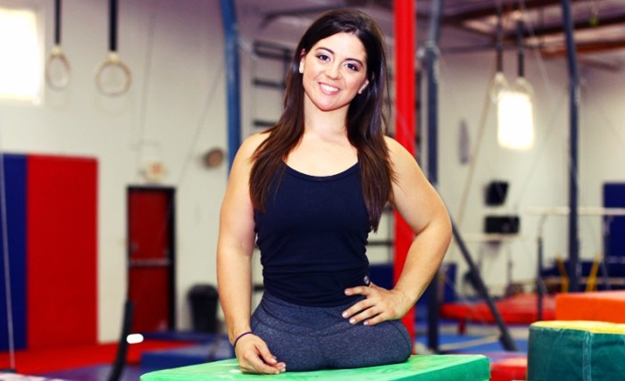 Con un gimnasio de fondo, Jennifer sonríe a cámara en camiseta y licra negra.