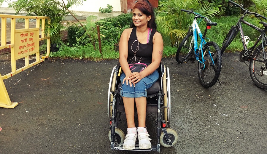Nisha sentada en su silla de ruedas, en camiseta, con unos audífonos en su cuello, sonríe coquetamente a la cámara 