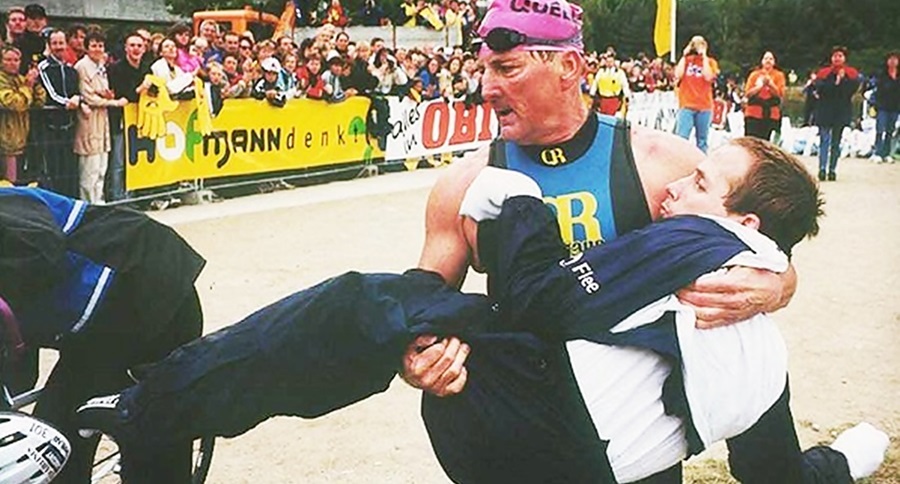 Dick, vestido con traje deportivo, carga en brazos a su hijo, también vistiendo ropa deportiva, en la meta de alguna de sus carreras, el público observa tras ellos. 