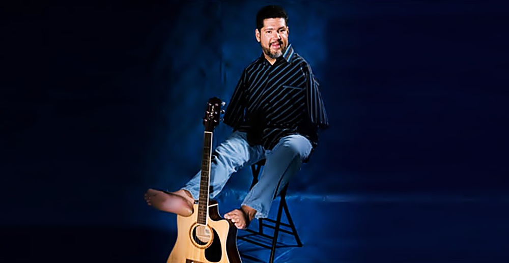 Sentado en un banco, con una medio sonrisa en su rostro con barba, Tony sostiene una guitarra