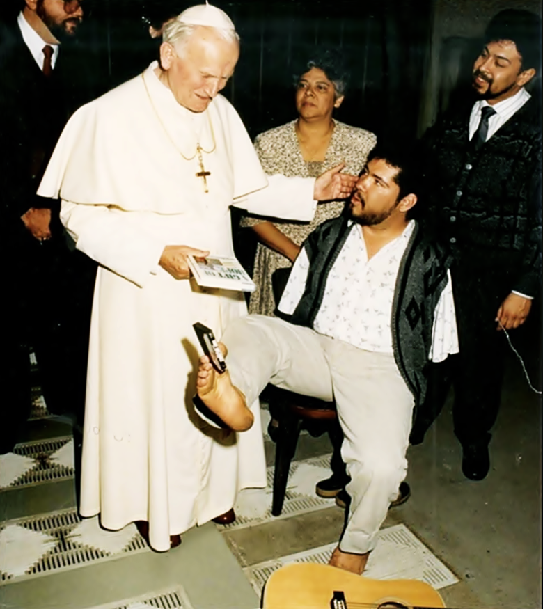 Juan Pablo segundo, parado, recibe presentes con los pies de Tony sentado en un banco