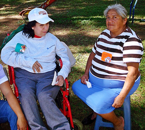 Bajo la sombra de un árbol, Sandra en su silla de ruedas y doña Marcelina en un banco de plástico. 