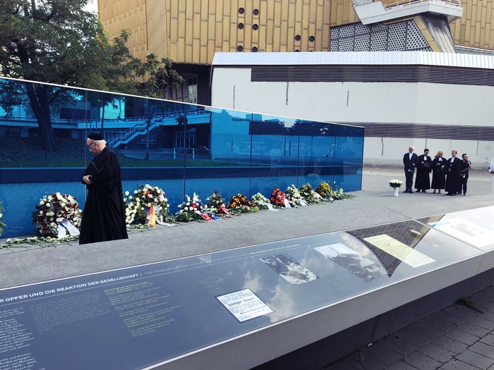 Un muro de cristal azul en una calle, en su base algunas ramos de flores y unas placas al otro lado de la calle. Se ve un sacerdote y varias personas con atuendo de jueces.