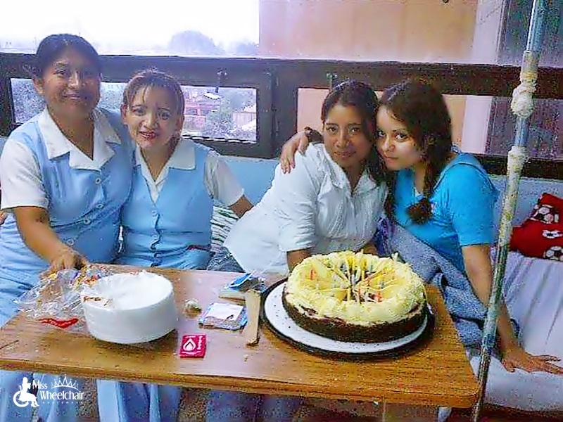 En la habitación de un hospital, 2 enfermeras sonríen sentadas en la cama de María quien sentada con los pies a lo largo, abraza a una amiga. Al frente está una mesa con un pastel con velas y platos de platicó. 