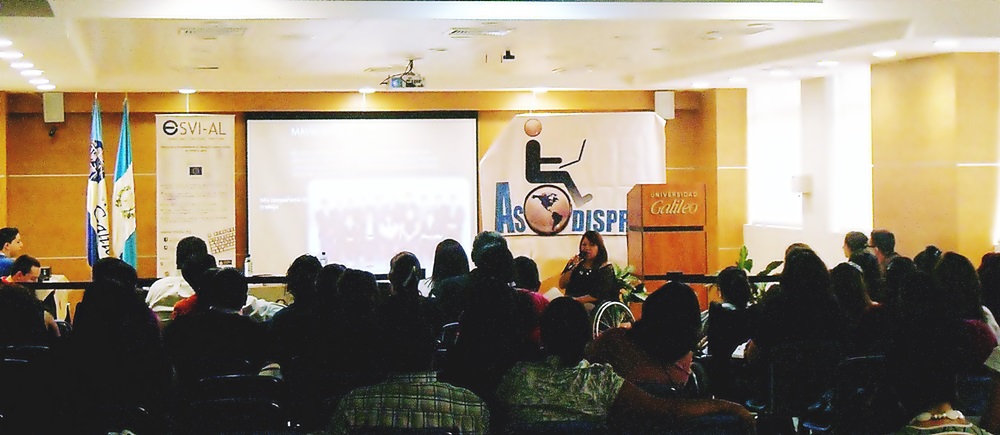 Fotografía del auditorio principal de Universidad Galileo, se aprecia lleno de personas y al frente, en el escenario, una mujer en silla de ruedas desarrolla una conferencia de ASODISPRO