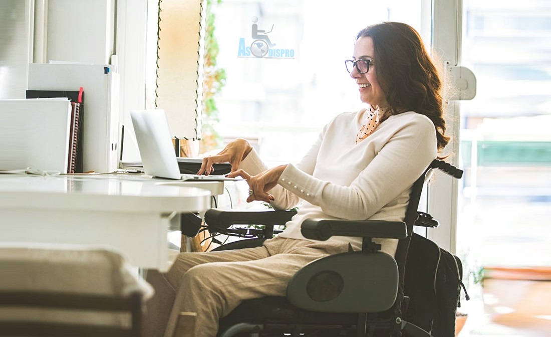 Una mujer de lentes, en silla de ruedas, trabaja en su computadora, en una oficina adornada con plantas. 