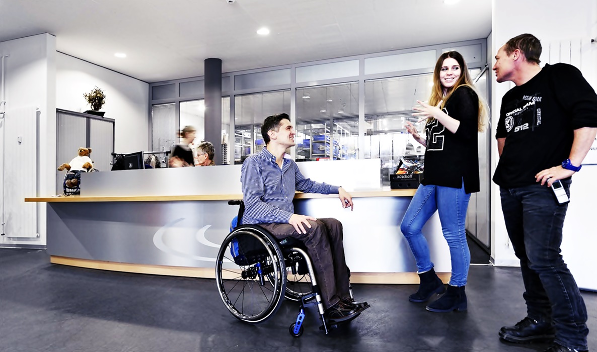 Frente al mostrador de una empresa, un joven en silla de ruedas conversa sonriente con una mujer y otro hombre, al parecer, compañeros de trabajo.