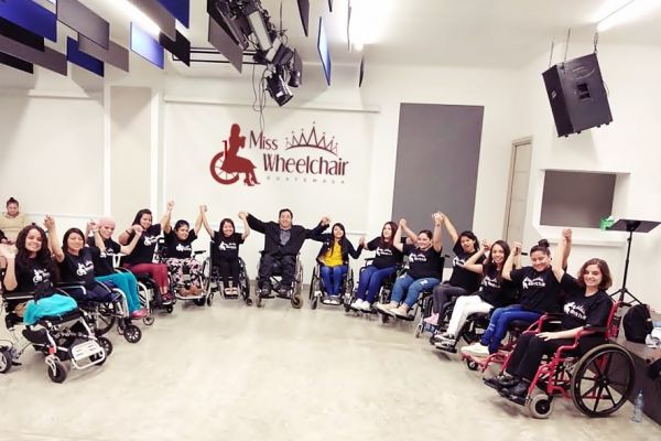 En un salón bajo reflectores, Byron Pernilla posa junto a las candidatas y directora de Miss Wheelchair Guatemala, todos hace una media luna con sus sillas de ruedas y se agarran las manos mientras las levantan.