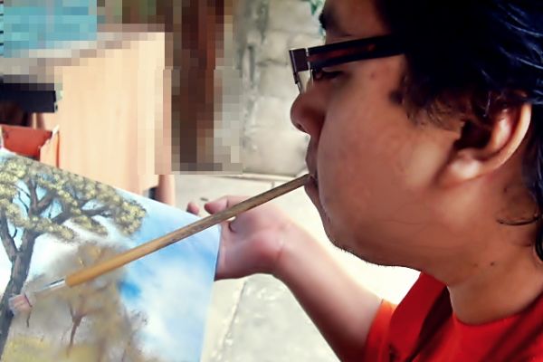 Byron Pernilla sostiene un pincel con la boca y pinta un árbol en un lienzo.