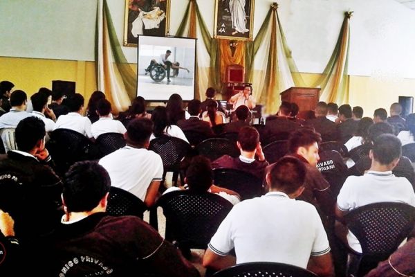 En un auditorio lleno de público, Byron Pernilla desarrolla una conferencia para estudiantes de nivel medio. Se observa a Byron con micrófono y a su lado una pantalla un una diapositiva de una persona con discapacidad bailando con otra sin discapacidad.