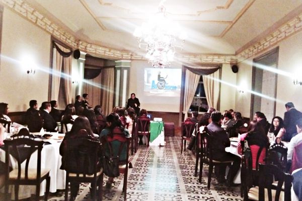 En un salón con iluminación nocturna, se observa un público sentado en varuias mesas, al fondo en un escenario, Byron Pernilla realiza una presentación con diapositivas.