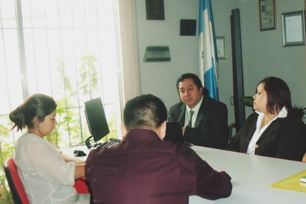 En una mesa de juntas con una bandera de Guatemala, Byron Pernilla da declaraciones a 2 reporteros de Prensa Libre, a su lado está Lissette Véliz.
