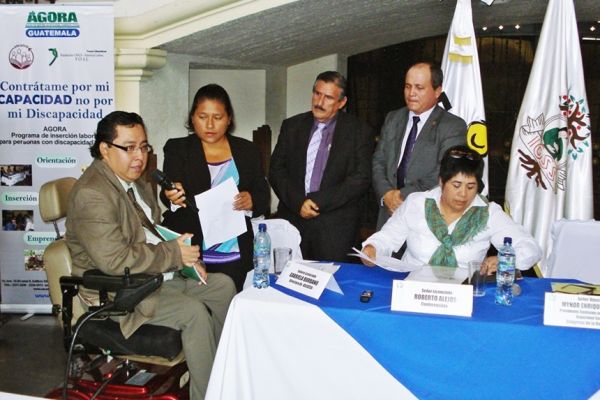 En un salón con varias banderas al fondo, Byron Pernilla de saco y corbata en su silla de ruedas, se dirige a varios diputados del Congreso de Guatemala durante la entrega de una iniciativa de ley en favor de personas con discapacidad.