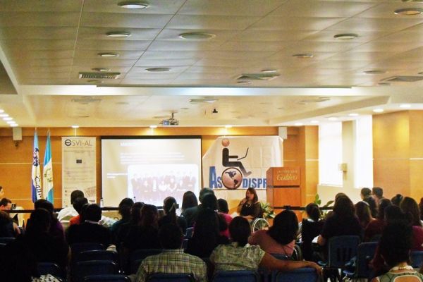 En el auditorio principal de Universidad Galileo, una capacitadora en silla de ruedas se dirige al público asistente.